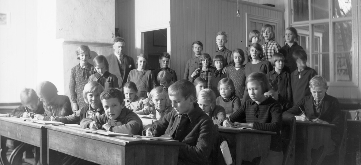 Gruppfoto av skolbarn med läraren i bakgrunden. Eleverna i full färd med att skriva något.